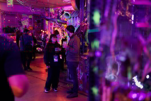 People are standing and talking in a colorful room with lights and decorations, holding drinks and event badges.