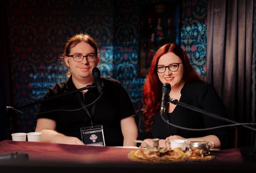 A pair of hosts sit at microphones in a cozy studio, smiling at the camera.