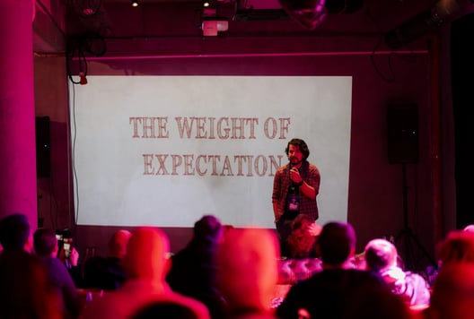 A speaker stands before a large slide titled “The Weight of Expectation,” addressing an attentive audience.