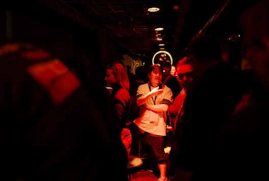 A person animatedly talks with others in a crowded, red-lit event space.