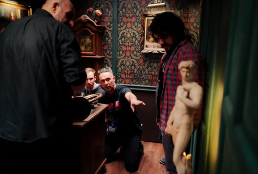 A participant kneels and points something out to the group inside a richly decorated escape-room setting.